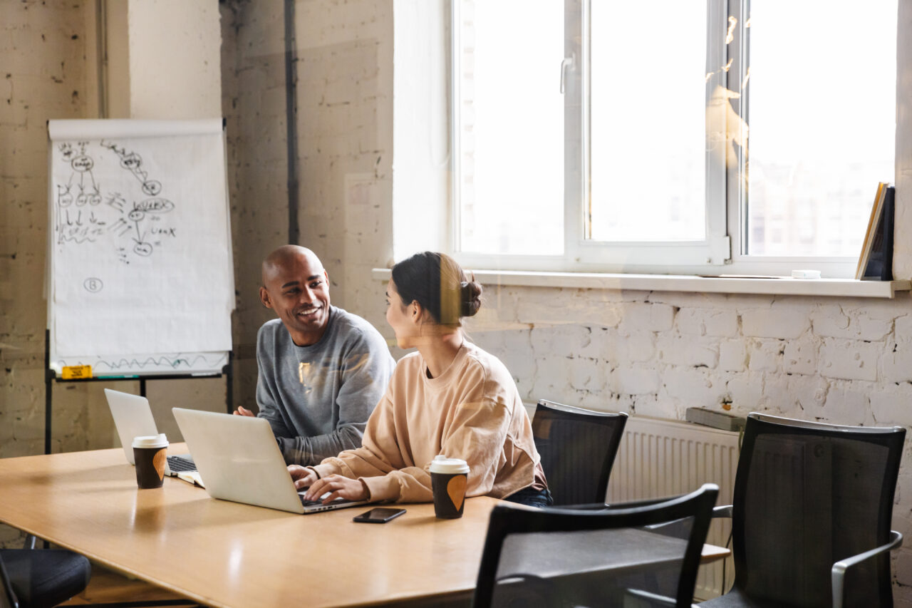 A man, business mentor, and a woman, a business owner, are sitting side by side during a business coaching session. They are talking to each other and appear to be in a happy mood.
