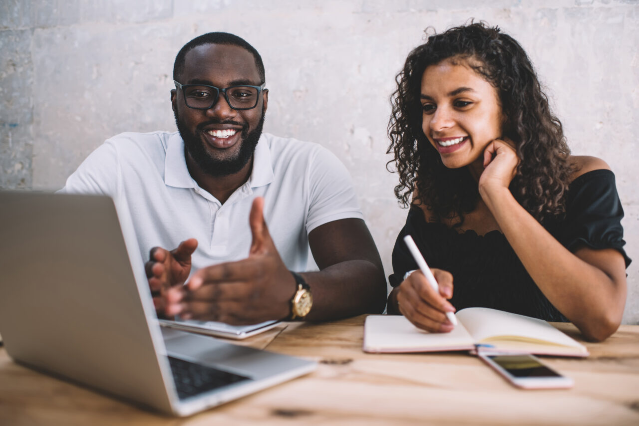 A business coach (man on the left) is showing available programs to a business owner (woman on the right) on his laptop. The mentor is explaining something to her.
