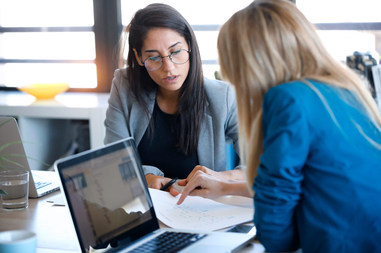 A female business owner is showing a document to the business mentor and asking a question. Both of them are deeply focused on the topic. They are examining the document while discussing issues.
