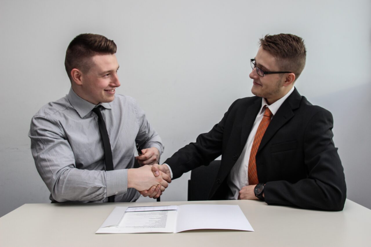 An man is shaking hands with another man while sitting down together. One of them is an HR personnel who seems to be welcoming the other one as a new in-house employee.